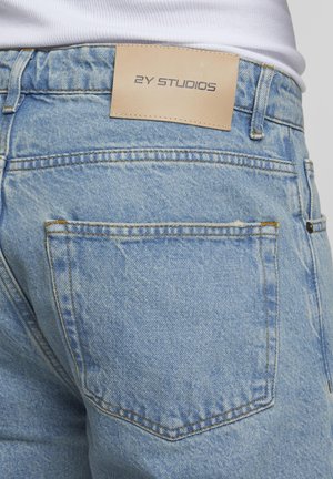 Lichtblauwe spijkerbroek achterkant met een leren patch met de tekst "2Y Studios" boven de rechterzak, gedragen met een wit overhemd.