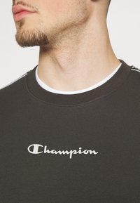 Primo piano del collo e del mento di un uomo che indossa un felpa scura con il logo "Champion" bianco centrato sul petto e un colletto di una maglietta bianca visibile.