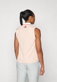 Růžový polo triko bez rukávů s bílým límcem, má na zadní straně červené logo adidas a postranní rozparky pro větší pohodlí.