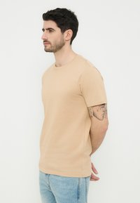 Άνδρας με σκούρα μαλλιά που φοράει μπεζ μπλουζάκι και ανοιχτόχρωμο μπλε τζιν, στέκεται προφίλ με τα χέρια πίσω από την πλάτη, δείχνοντας ένα τατουάζ στο αριστερό του χέρι.