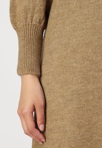 Pull en maille beige chaud, avec des poignets côtelés et une texture lisse. Une main à la couleur de la peau repose doucement sur le tissu.