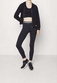 Abbigliamento sportivo nero composto da una felpa cropped con zip, leggings aderenti e un reggiseno sportivo. Tessuto testurizzato con una cintura a contrasto. Sneakers nere.