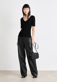 Črn rebrast pleten bluzon z V-izrezom, v kombinaciji z visokimi črnimi usnjenimi širokimi hlačami. Manekenka drži malo črno torbico.