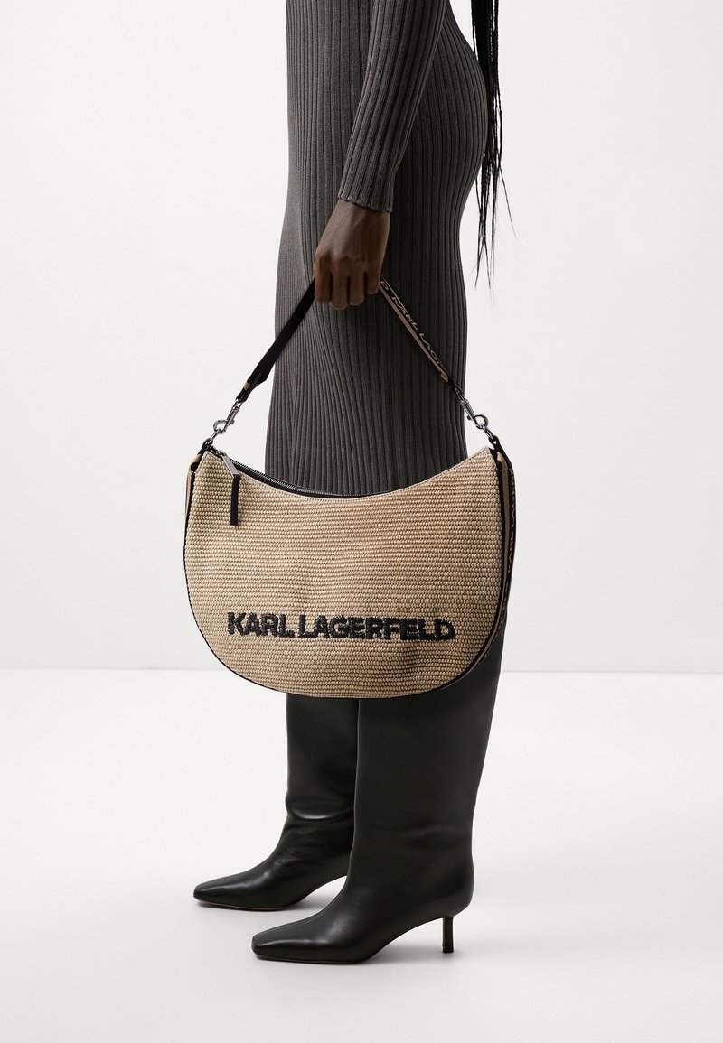 Vue de profil d'une personne en robe côtelée et bottes noires hautes jusqu'au genou, tenant un sac à main tissé beige Karl Lagerfeld avec une bandoulière noire.