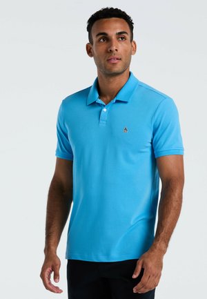 Homme portant un polo à manches courtes bleu vif avec un petit logo de pingouin, se tenant devant un fond clair, regardant légèrement à droite.