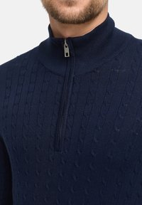 Granatowy sweter wełniany z wysokim kołnierzem i półzamkiem. Posiada teksturowany wzór w warkocze na całej powierzchni materiału.