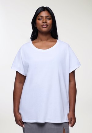 T-shirt bianco in cotone con maniche corte e scollatura larga a giro. Vestibilità comoda con una superficie liscia, indossato sopra una gonna grigia con spacco laterale.
