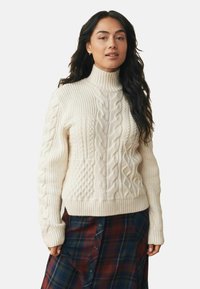 Cream cable knit turtleneck sweater with textured patterns, ribbed cuffs, and hem, paired with a plaid skirt in dark blue, red, and green.