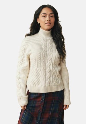 Cream cable knit turtleneck sweater with textured patterns, ribbed cuffs, and hem, paired with a plaid skirt in dark blue, red, and green.