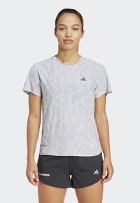 Mujer vestida con una camiseta atlética gris claro con estampado y pantalones cortos negros con logotipos, de pie contra un fondo liso claro.