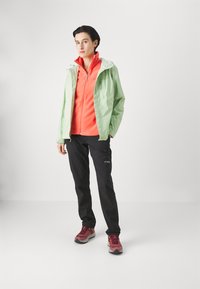 Giacca impermeabile verde chiaro con cappuccio sopra un felpa arancione brillante, pantaloni neri e scarpe sportive bordeaux.