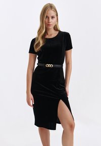 Czarna aksamitna sukienka z okrągłym dekoltem, krótkimi rękawami i wycięciem do uda. Uzupełniona czarnym paskiem z złotą klamrą.