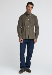 Olivgrön långärmad skjorta med knappnerkoll, kombinerad med blå denimjeans och mörka skor med orange detaljer.