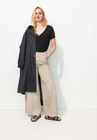 Body noir à côtes, pantalon large beige, et un long manteau noir. Sandales à lanières noires. Texture lisse, avec des boutons et des poches visibles.