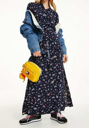 Marineblauwe maxi-jurk met bloemmotief en korte mouwen, een tailleriem, een gebruikte spijkerjas, een gele crossbodytas en zwarte sneakers met rode accenten.