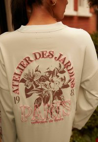 Krémszínű pulóver, amely grafikus virágmintázattal rendelkezik, pink betűkkel "ATELIER DES JARDINS 1983" és "PARIS collection de fleurs" felirattal.