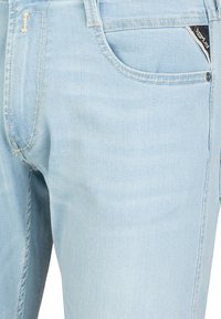 Helle blaue Denim-Jeans mit glatter Textur, ausgestattet mit einer Vordertasche, sichtbaren Nähten und einem kleinen schwarzen Etikett in der Nähe der Tasche.