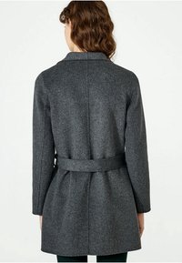 Žena oblečená v šedém vlněném kabátu střední délky s páskem uvázaným v pase, zobrazená zezadu na jednobarevném pozadí.