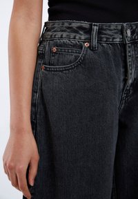 Högmidjade svarta jeans i denim med raka ben, prydda med kopparnitar, sidofickor och en fransig midja. Ytan är något sliten.