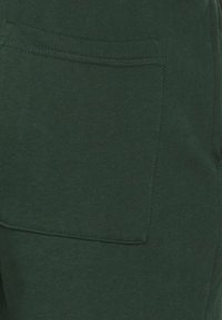 Tmavě zelené bavlněné kalhoty se střihem rovného střihu, vybavené jednou náprsní kapsou na boku. Materiál má hladkou a měkkou texturu.