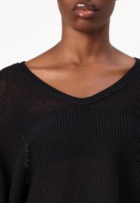 Pull noir en tricot avec un col en V, présentant une texture à mailles ouvertes et une coupe décontractée, mettant en valeur le décolleté et la zone des épaules.