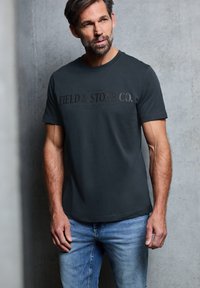Tmavě šedé bavlněné tričko s krátkým rukávem, s vyraženým nápisem "FIELD & STONE CO." přes hrudník. Nošeno s světle modrými džínami.