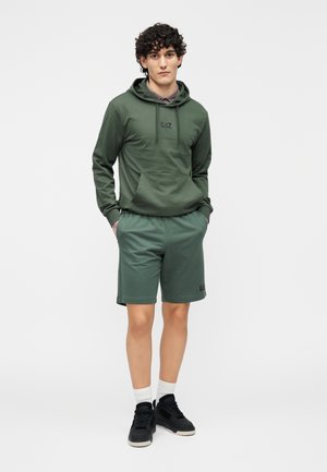 Φούτερ με κουκούλα σε ελιά πράσινο χρώμα με εμπρόσθια τσέπη και λογότυπο, συνδυασμένο με ταιριαστά πράσινα σορτς. Μαύρα αθλητικά παπούτσια και λευκές κάλτσες ολοκληρώνουν την εμφάνιση.
