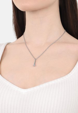 Collana in argento con pendente a goccia che presenta una piccola pietra preziosa trasparente, abbinata a una blusa a righe chiara.
