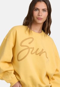 Sweatshirt jaune en coton doux avec un grand motif "Sun" en cursive de couleur marron clair. Coupe ample avec des poignets côtelés.
