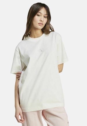 Junge Frau mit langen dunklen Haaren, die ein lockeres weißes T-Shirt mit kurzen Ärmeln und eine hellrosa Hose trägt und vor einem einfarbigen Hintergrund posiert.