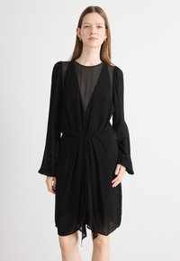 Rochie neagră cu decolteu în V și mâneci lungi din material transparent, având un tecido texturat și o talie strânsă. Se lasă ușor până la mijlocul coapsei.
