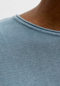 Gros plan sur une épaule à peau foncée portant une chemise en tissu côtelé bleu clair avec des coutures visibles au niveau de l'encolure.