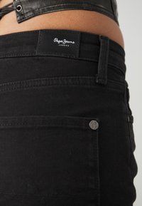 Jeans neri con etichetta "Pepe Jeans London", materiale in denim, cuciture a contrasto e un motivo a onda decorativo sulla tasca posteriore.