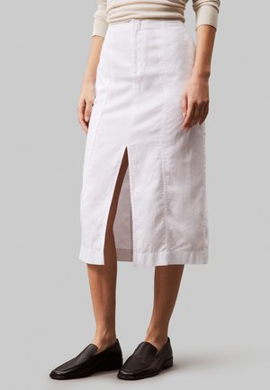 Witte katoenen midi-jurk met een split aan de voorkant, rechte snit, subtiele textuur en nette stiksels. Gepaard met zwarte instap schoenen.