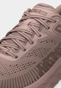 Κοντινό πλάνο ενός αθλητικού παπουτσιού Hoka One One σε ροζ χρώμα με υφή πλεκτού στο πάνω μέρος και σφιχτά δεμένα επίπεδα κορδόνια.