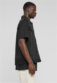 Chaleco de mezclilla negra con un diseño clásico, que cuenta con dos bolsillos en el pecho, botones a presión y un cuello con solapa. Lleva puesto un shirt negro.