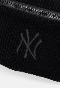 Svart kordfløyelsstoff med ribbet tekstur, med en svart brodert New York Yankees-logo og en svart glidelåsdetalj.