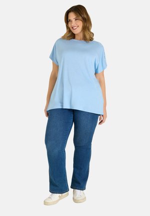 Frau lächelt, trägt ein lockeres, hellblaues Kurzarmshirt, blaue Jeans und weiße Turnschuhe, steht vor einem einfarbigen Hintergrund.