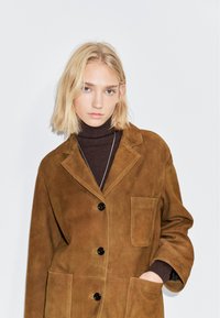 Giacca di camoscio marrone con colletto, tasca anteriore e tre bottoni neri. Indossata sopra un dolcevita marrone scuro, caratterizzato da una texture liscia.