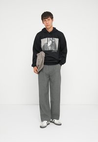 Schwarzer Hoodie mit Grafikdruck, graue Jogginghose und weiße Sneaker mit schwarzen Streifen. Das Model trägt eine hellgraue Tasche über einer Schulter.