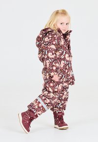 Kinderen winteroverall in bordeauxrood met een bloemen- en dierenpatroon. Gevoerd met een capuchon en reflecterende accenten. Gecombineerd met bruine winterlaarzen.