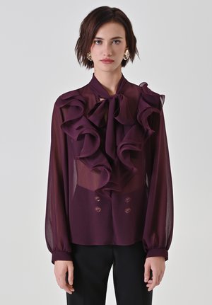 Giovane donna che indossa una blusa di plissettata prugna trasparente con ampie balze e un fiocco al collo, abbinata a pantaloni neri, in piedi di fronte a uno sfondo semplice.