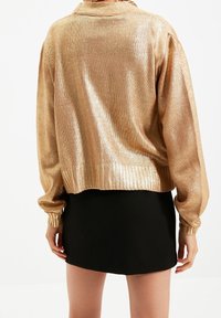 Goldfarbener, metallischer, kurz geschnittener Pullover mit gerippten Bündchen und Saum, mit lockerer Passform und strukturierter Oberfläche. Kombiniert mit einem schwarzen Minirock.