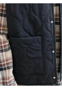 Marineblå quiltet vest med bølget mønster, der har en sidelomme og trykknaplukninger, lagdelt over en ternet flannel skjorte i beige og brune nuancer.