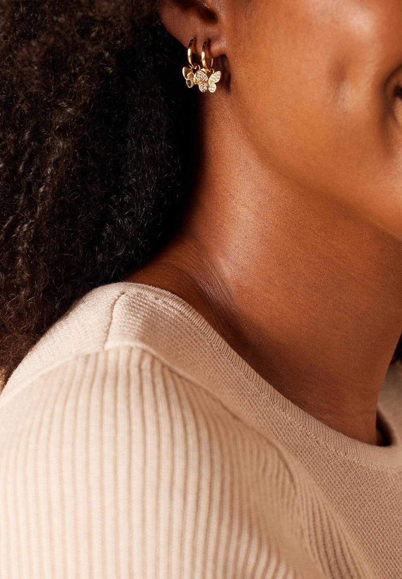 Goldene Creolenohrringe mit Schmetterlingsanhängern, die mit klaren Strasssteinen besetzt sind, vor einem strukturierten, hellbeigen Pulloverkragen.