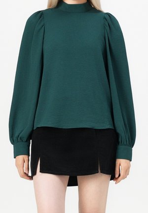 Kobieta ubrana w ciemnozieloną, długorękawową, bufiaste bluzkę oraz czarną mini spódniczkę z przednimi rozcięciami, stojąca na jednolitym tle.