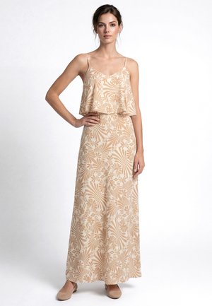 Nő bézs mintás, ujjatlan maxiruhában és lapos cipőben, egy kezét csípőjén tartva pózol fehér háttér előtt.