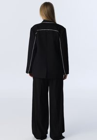 Zwarte op maat gemaakte blazer met witte stikdetails, gecombineerd met zwarte wijde broek. Soepele textuur, gestructureerd ontwerp en moderne lijnen.