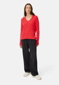 Roter gerippter Strickpullover mit V-Ausschnitt, kombiniert mit lockeren schwarzen Hosen und weißen Sneakers. Die Textur wirkt weich und lässig.