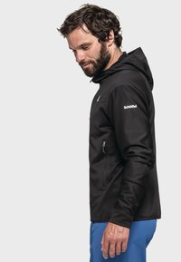 Schwarze Kapuzenjacke mit einer Reißverschlusstasche. Das Material wirkt weich und hat eine glatte Textur. Logo auf dem rechten Ärmel. Modell trägt blaue Hosen.
